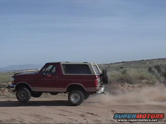 Red Ford Bronco with a tan top, kicking up dust on a dirt road. Spare tire mounted on the rear