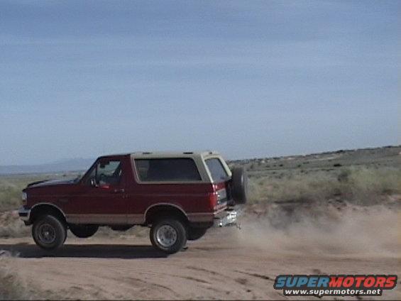 Red 1994 Ford Bronco driving on a dirt road, kicking up dust. Spare tire mounted on the back