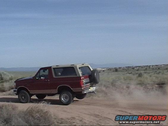 Red 1994 Ford Bronco driving on a dirt road, kicking up dust. Spare tire on the back