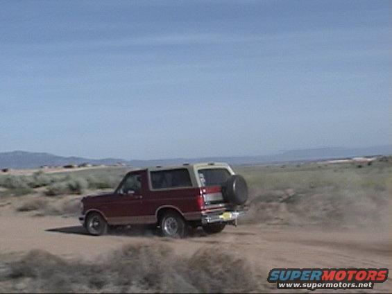 Red and tan Ford Bronco with a spare tire on the back, driving on a dirt road