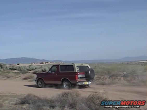 Maroon 1994 Ford Bronco with a tan top driving on a dirt road, kicking up dust. Spare tire mounted on the rear