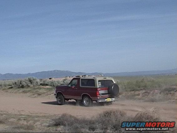 Red and tan 1994 Ford Bronco driving on dirt road, rear view