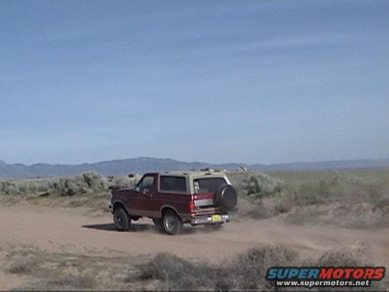 Red and tan Ford Bronco with a spare tire on the back, driving on a dirt road