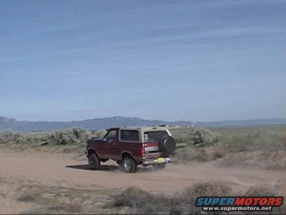 Red and tan 1994 Ford Bronco driving on dirt road, rear view