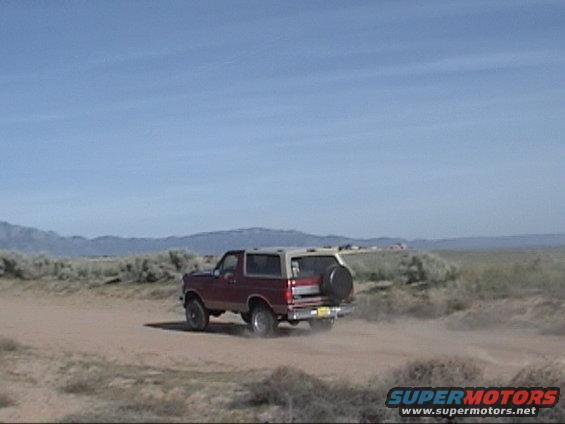 1994 Ford Bronco driving on a dirt road, kicking up dust. Spare tire mounted on the rear