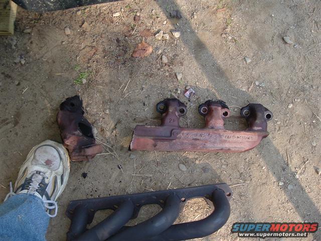Two rusty and one black exhaust manifold on dirt ground next to a shoe