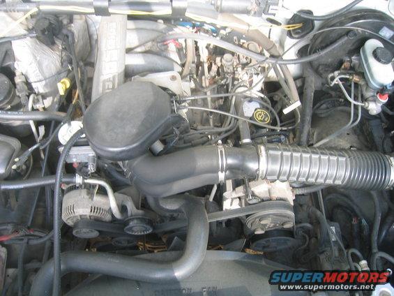 Engine bay with hoses, belts, and alternator visible. Some text on a fan is partially obscured