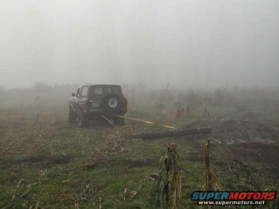 Dark 1978 Ford Bronco with a spare tire on the rear, pulling a log in a foggy field
