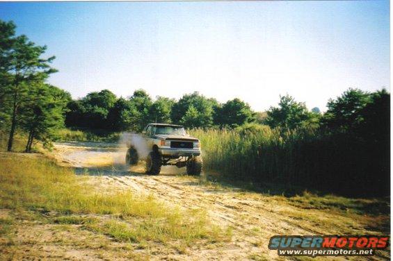 Pickup truck kicking up dust on a dirt road, trees and tall grass in background
