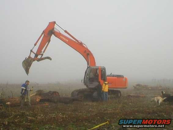 Orange excavator with two people and a dog in a foggy field with logs