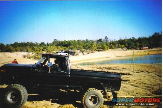 Black 1979 Ford F-250 pickup truck with large tires parked on sandy terrain near a body of water. Other vehicles and people are visible in the background