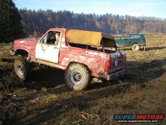 Red Bronco with tan soft top and large tires in mud, green SUV in background