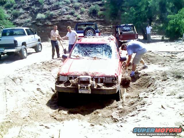 People trying to dig out a red off-road vehicle stuck in a dirt pit. Another truck is parked nearby