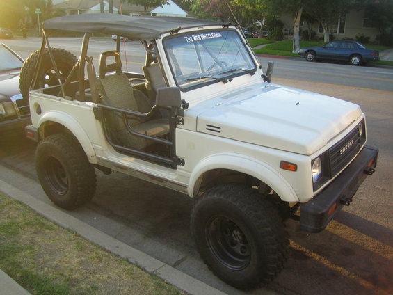 White Suzuki Samurai parked on street, soft top up, spare tire on back. "The Web Wheeler.com" sticker on windshield