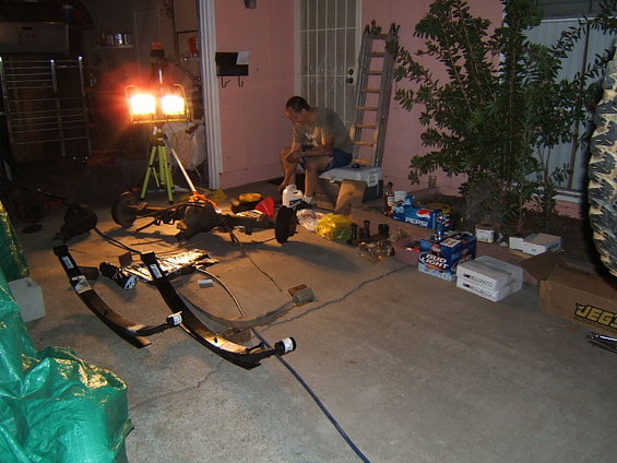 Vehicle axle and leaf springs laid out on concrete, with a man sitting nearby and work lights on