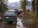 1978 Ford Bronco - Several off-road vehicles navigate a wet, wooded trail with mountains in the background.