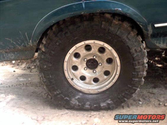Dark green SUV's front wheel with a silver multi-hole rim and aggressive tire, mud splattered on the fender