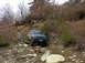 1978 Ford Bronco - A blue SUV is seen off-roading on a steep, rocky incline surrounded by trees and brush.