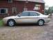 2000 Ford Taurus - A gold 2000 Ford Taurus sedan is parked on a gravel lot in front of a red wooden building with signs.