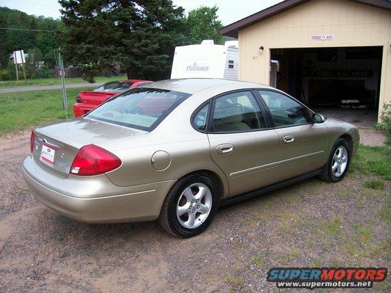 Gold 2000 Ford Taurus sedan parked on gravel, rear 3/4 view. A red sports car is visible behind it