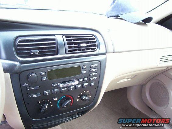 Interior view of a 2000 Ford Taurus dashboard showing the radio, air vents, and climate controls. A blue baseball cap rests on the passenger side