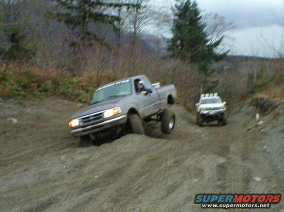 Two lifted trucks driving on a dirt hill, one in the foreground and one further back