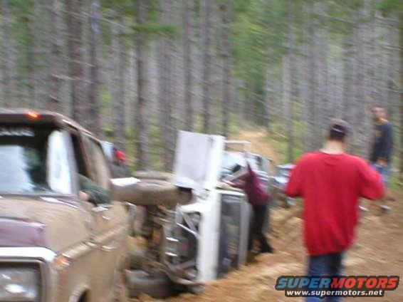 Overturned 1978 Ford Bronco and another vehicle in a forest clearing with two people nearby