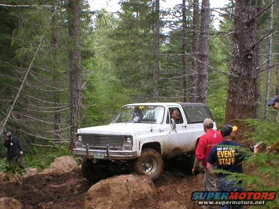 White 1975 Chevrolet K5 Blazer off-roading on a muddy trail with trees in the background