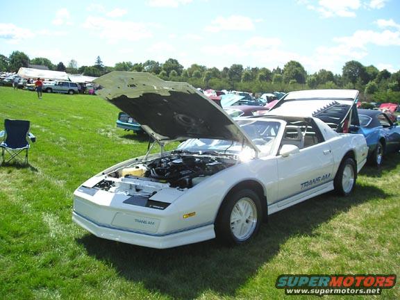 White 1984 Pontiac Trans Am with hood and trunk open, parked on grass at an outdoor car show