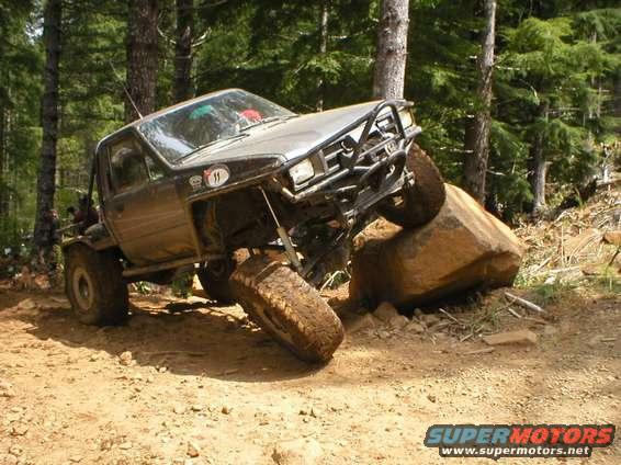 Off-road truck with large tires climbing over rocks on a dirt trail. It has a custom front bumper and roll cage