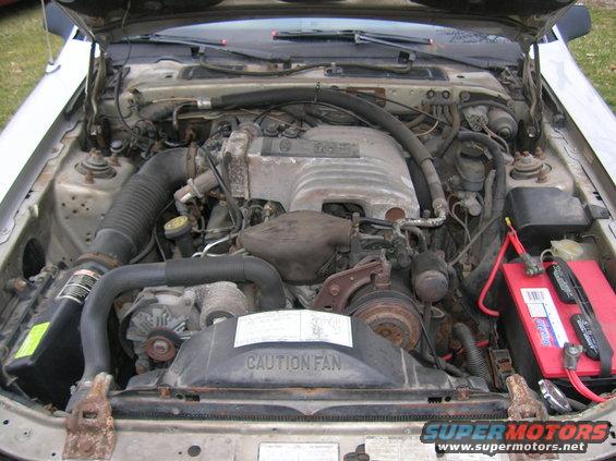 Car engine bay with V6 engine, battery, and hoses visible. Caution Fan label on shroud