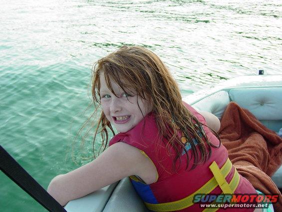 Girl with wet hair and red life jacket smiles from a boat on the water