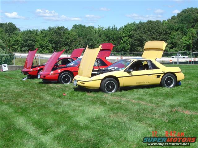 Three 1984 Pontiac Fieros parked on grass, hoods and rear decks open. Yellow Fiero in foreground has 'Fiero' script on side