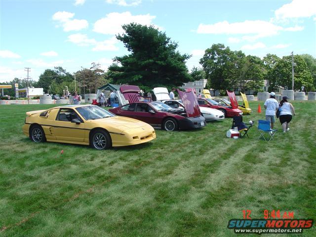 Yellow 1984 Pontiac Fiero in front of other Fieros on grass, with people and tents in background
