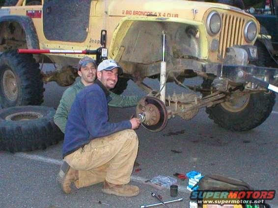 Two men working on the rear axle of a yellow Jeep CJ, with tools and spare tires nearby. Text on the Jeep reads '2 BIG BRONCOS 4 X 4 CLUB'