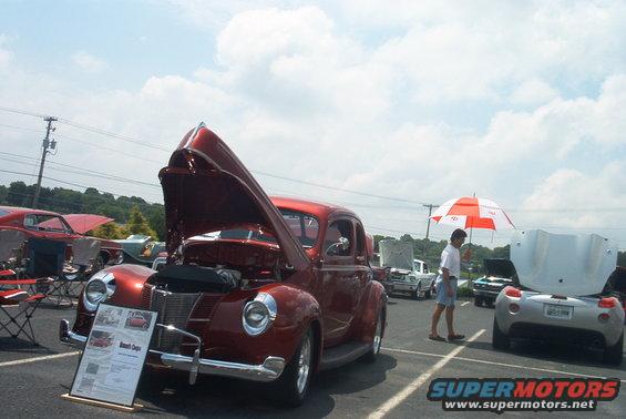 Red 1940s coupe with hood open at a car show, display board in front. Other cars and people visible