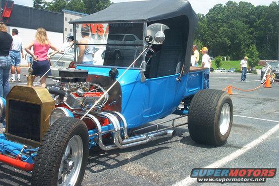 Blue hot rod with a large V8 engine, chrome exhaust, and wide rear tires. Black soft top is raised