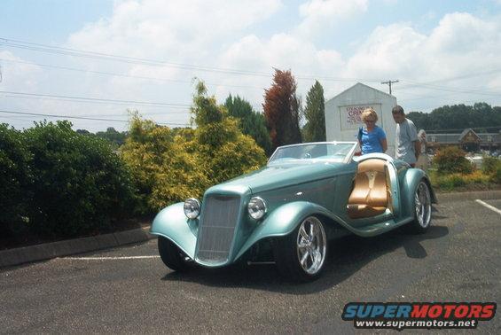 Teal custom hot rod convertible with tan interior parked in a lot, two people standing nearby