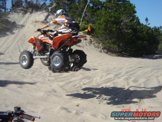 Quad bike mid-air over sand dune, rider wearing helmet and orange/white gear. Trees and blue sky in background