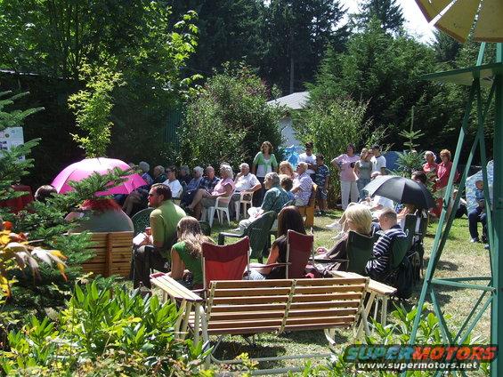 People gathered outside on a sunny day, sitting in chairs and on a bench, with trees and a house in the background