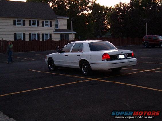 White sedan parked in a lot, rear view. Police light on roof. Red SUV in background