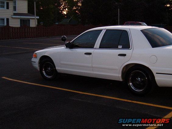 White sedan parked in a lot, side view. The car has black wheels and a spotlight on the driver's side