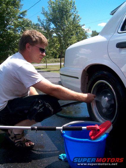 Guy washing a white car's wheel with a hose and brush. Blue bucket nearby