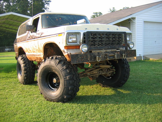Mud-splattered white and brown Ford Bronco with lifted suspension and large off-road tires, parked on grass in front of a white garage