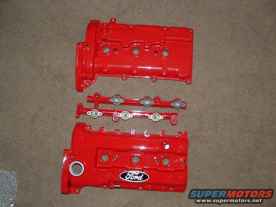 Two red Ford valve covers and fuel rails on a carpeted surface. The Ford logo is visible on one valve cover