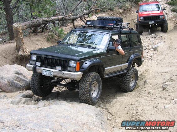 Green 1996 Jeep Cherokee with off-road tires and roof rack navigating a rocky trail, red Jeep Cherokee behind it