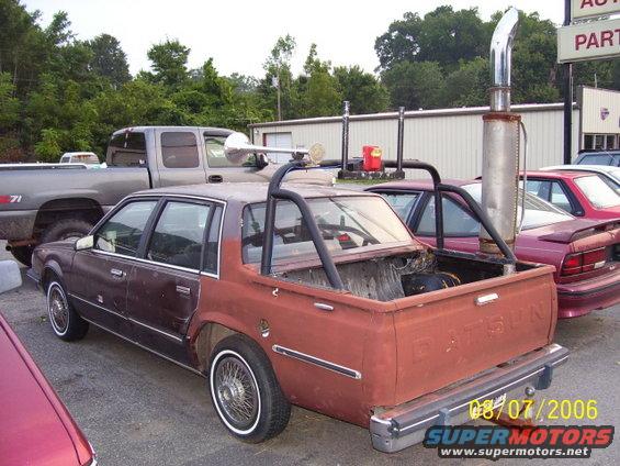 Custom Datsun pickup with roll cage and large exhaust stack parked next to a pickup truck and other cars