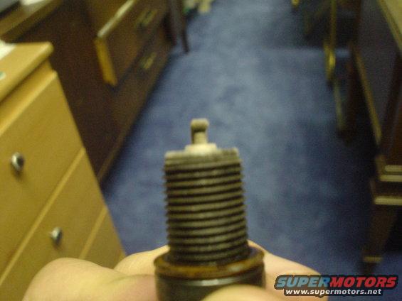 Spark plug held in hand, showing threads and electrode. Background is blurred