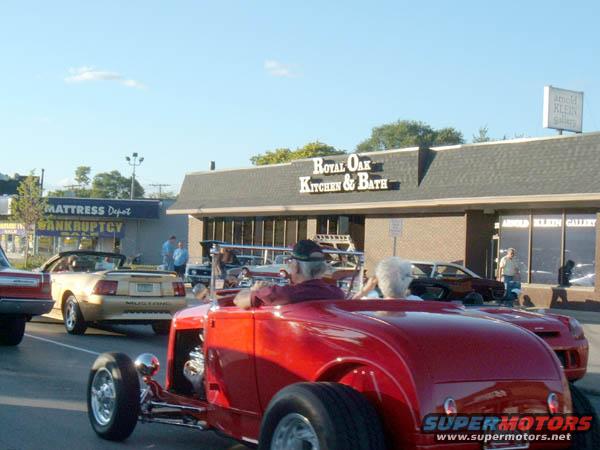Red hot rod in foreground, gold convertible Mustang behind it, and other cars on street in front of businesses