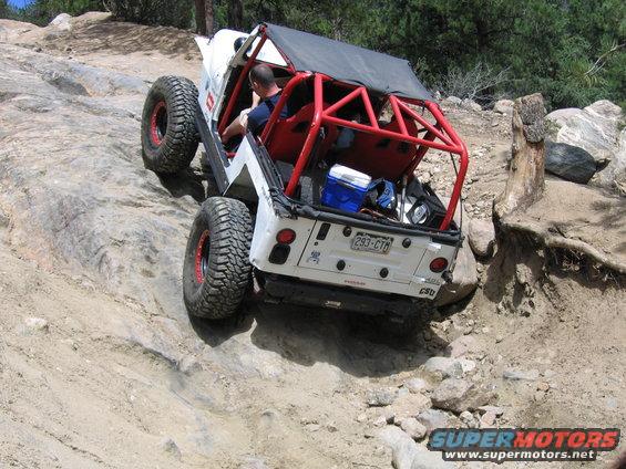 Jeep Wrangler climbing a rocky hill, driver visible, red roll cage, cooler in back